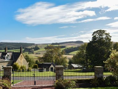 View of a gated entrance with stone pillars leading to houses and rolling hills in the background at The Lodge (Elsdon) in Elsdon