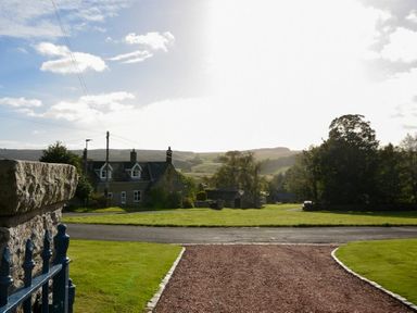 A gravel driveway leading to a road with a stone house and green fields in the background at The Lodge (Elsdon) in Elsdon