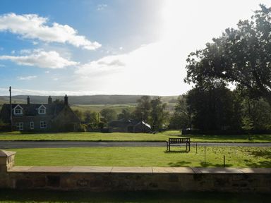 A grassy yard with a wooden bench and stone wall with a house and trees in the background at Rose Cottage Elsdon