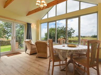 A dining room with a round table covered with a tablecloth and wooden chairs near large windows overlooking a garden at Rose Cottage Elsdon