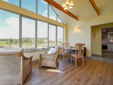 A dining area with wooden chairs and table next to wicker armchairs and large windows showing a countryside view at Rose Cottage Elsdon