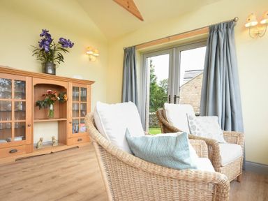 A sitting area with two wicker chairs facing glass doors and a wooden cabinet with flowers at Rose Cottage Elsdon