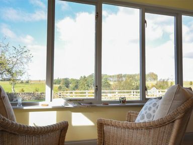 Two wicker chairs with cushions by a large window overlooking a grassy landscape at Rose Cottage Elsdon