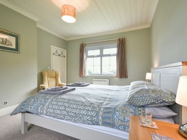 A bedroom with a bed and patterned bedding a beige armchair and a window with curtains at Rose Cottage Elsdon