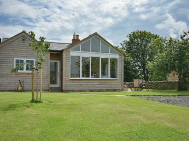 A wooden house with large windows and a green lawn with trees and outdoor chairs at Rose Cottage Elsdon