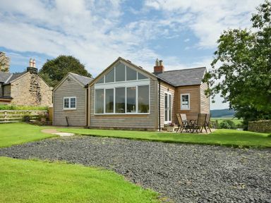 A small wooden house with large windows and outdoor chairs on grass at Rose Cottage Elsdon