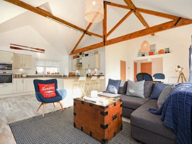An open living space with a gray sectional sofa a wooden trunk coffee table a blue armchair with a red cushion a kitchen with bar stools and exposed wooden ceiling beams at Bowline Cottage in Beadnell