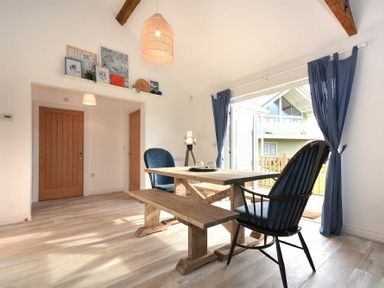 A dining area with wooden table bench and chairs near large windows with blue curtains at Bowline Cottage in Beadnell