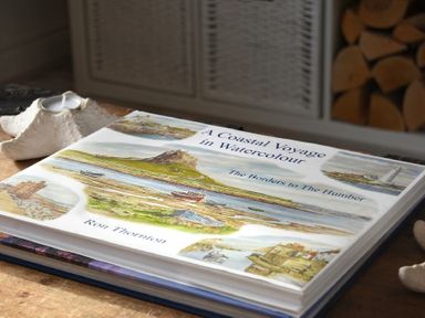 A watercolor art book titled A Coastal Voyage in Watercolour on a wooden table with a shell candle holder nearby at Bowline Cottage in Beadnell
