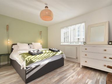 A bedroom with a bed, pillows, green throw, bedside lights, window with blinds, white dresser with a mirror and clock at Bowline Cottage in Beadnell