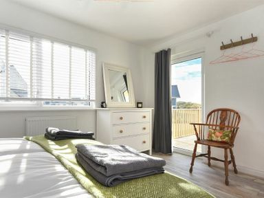 A bedroom with a bed, chest of drawers with mirror, wooden chair, and door leading to a balcony at Bowline Cottage in Beadnell