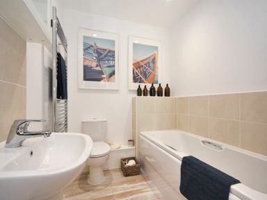 A bathroom with a sink, toilet, bathtub, two framed pictures, and bottles on the bathtub ledge at Bowline Cottage in Beadnell