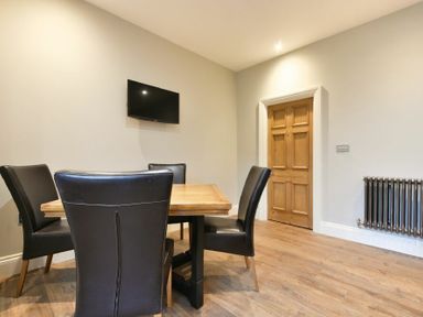 A dining area with a wooden table, black chairs, a wall-mounted TV, a wooden door, and a radiator at Sandybrae in Alnmouth