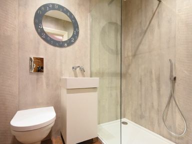 A small bathroom with a wall-mounted toilet a white sink a glass shower enclosure and a circular mirror with numbers on the frame at Sandybrae in Alnmouth