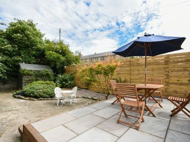 An outdoor patio with wooden table and chairs and a blue umbrella next to a small white table and chairs with greenery and a wooden fence in the background at Sandybrae in Alnmouth