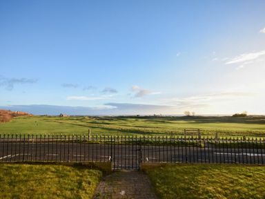 A fenced grassy garden area with a paved path overlooking a green field under a blue sky at Mariners House in Alnmouth
