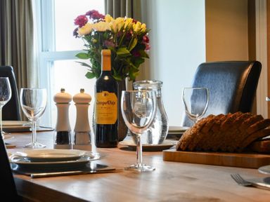 A dining table set with wine glasses plates a bottle of wine salt and pepper shakers flowers and sliced bread at Mariners House in Alnmouth