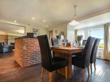 A dining area with wooden table and black chairs next to a brick half-wall and windows with curtains at Mariners House in Alnmouth