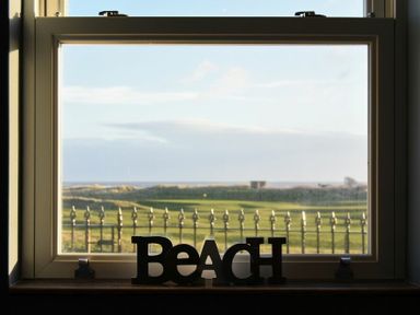 A window with a decorative beach sign on the sill overlooking a grassy area with a fence and horizon at Mariners House in Alnmouth