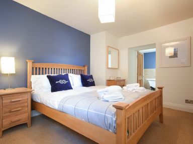 A bedroom with a wooden bed blue cushions bedside table lamp and an open door to a bathroom at Mariners House in Alnmouth