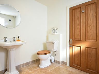 A bathroom with a pedestal sink a round mirror a toilet with a wooden seat and a wooden door at Mariners House in Alnmouth