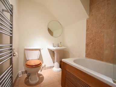A bathroom with a toilet, pedestal sink with round mirror, and a bathtub with wooden paneling at Mariners House in Alnmouth
