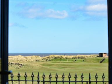 A view of a green golf course with sand dunes and the sea in the background seen through a window with a black iron railing