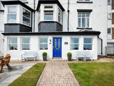 Front view of a white house with a blue door two windows on each side benches and potted plants on a brick pathway at Mariners House in Alnmouth