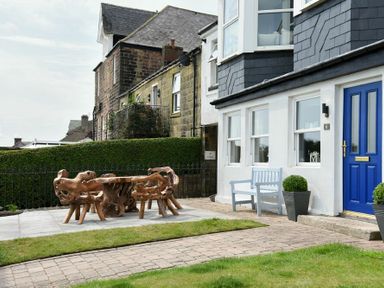 An outdoor patio area with wooden furniture and a blue door on a white building at Mariners House in Alnmouth