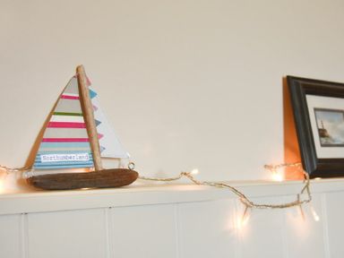 A wooden sailboat decoration with colorful striped sails and the word Northumberland alongside string lights on a white shelf near a framed picture at Farne View Cottage in Seahouses