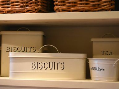 Metal storage containers labeled biscuits tea and nibbles on a shelf with wicker baskets above