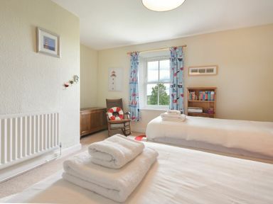 A bedroom with two beds white towels on a bed a wooden rocking chair a window with blue curtains and a bookshelf at Farne View Cottage in Seahouses