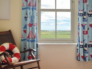 A window with blue curtains with boat patterns and a wooden rocking chair with a red and white life preserver pillow at Farne View Cottage in Seahouses