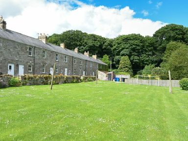 A row of stone houses with a grassy yard and trees in the background at Farne View Cottage in Seahouses