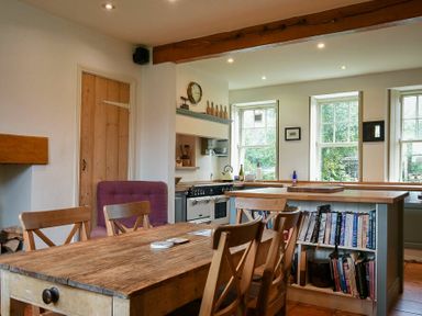 A kitchen with wooden dining table and chairs wooden countertop and bookshelf beneath windows at Whistlewood in Bamburgh