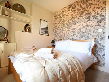 A bedroom with a wooden bed with white pillows and beige comforter with towels on top floral wallpaper and shelves with decorative items at Whistlewood in Bamburgh