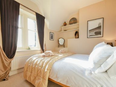 A bedroom with a bed covered with white sheets and a beige quilt near a window with curtains and a small dresser with a mirror and shelves with decorative items at Whistlewood in Bamburgh