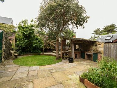 A garden with stone patio green grass trees outdoor bench and built in barbecue area at Whistlewood in Bamburgh