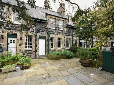 A stone house with multiple windows and white doors with a paved courtyard and various plants and trees at Whistlewood in Bamburgh