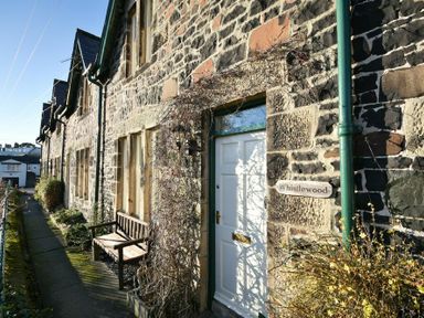 A stone exterior of a house with a white door and wooden bench near the entrance at Whistlewood in Bamburgh
