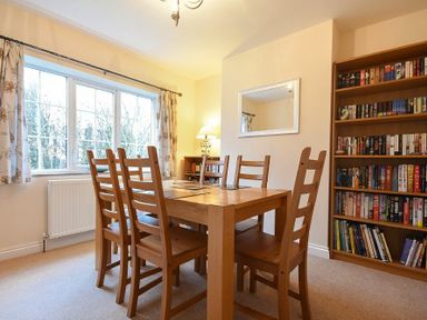 A dining room with a wooden table and six chairs a bookshelf filled with books a large window with curtains and a mirror on the wall at Castle View Warkworth