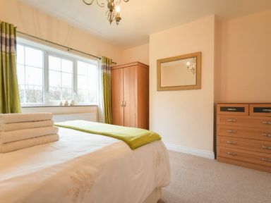 A bedroom with a bed covered in white sheets and green throw with a stack of towels on top next to a window with green and beige curtains and wooden wardrobe and chest of drawers with a mirror on the wall at Castle View in Warkworth