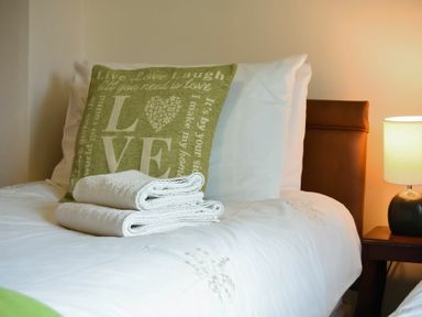 A bed with a green love-themed pillow folded white towels and a bedside table with a lamp at Castle View in Warkworth