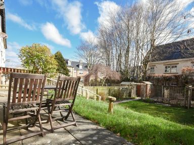 A patio with wooden chairs and a table next to a grassy garden with trees and houses in the background at Castle View in Warkworth