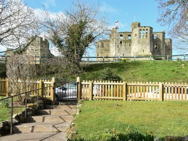 A stone castle on a hill behind a wooden fence and a garden with a gate and steps at Castle View Warkworth