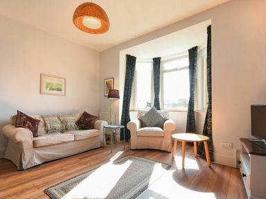 A living room with a sofa, armchair, lamp, two small tables, rug, TV, and bay windows with dark curtains at Rosedale House in Amble
