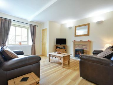 A living room with two leather sofas a wooden coffee table a TV on a wooden stand a fireplace and a window with curtains at Dryden Cottage in Bamburgh