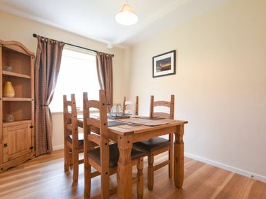 A dining room with a wooden table and chairs a cabinet with shelves a window with curtains and a framed picture on the wall at Dryden Cottage in Bamburgh