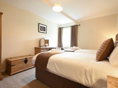 A bedroom with two single beds a wooden chest a dressing table and a window with curtains at Dryden Cottage in Bamburgh
