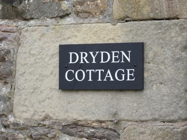 A black sign with white letters reading Dryden Cottage on a stone wall at Dryden Cottage Bamburgh